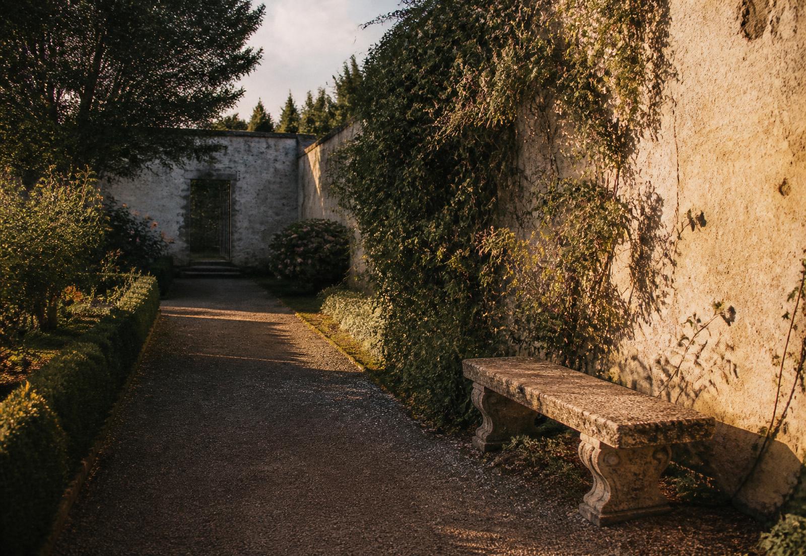 A quiet walled garden path at golden hour with a weathered stone bench